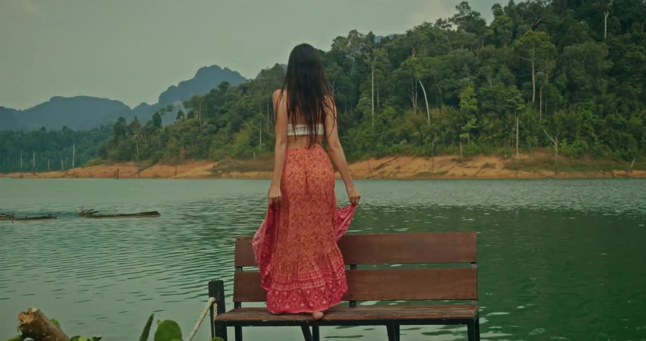 A carefree woman with long hair walks in slow motion on a wooden dock over a lake, smiling as she enjoys the beautiful natural scenery of the surrounding forest and mountains