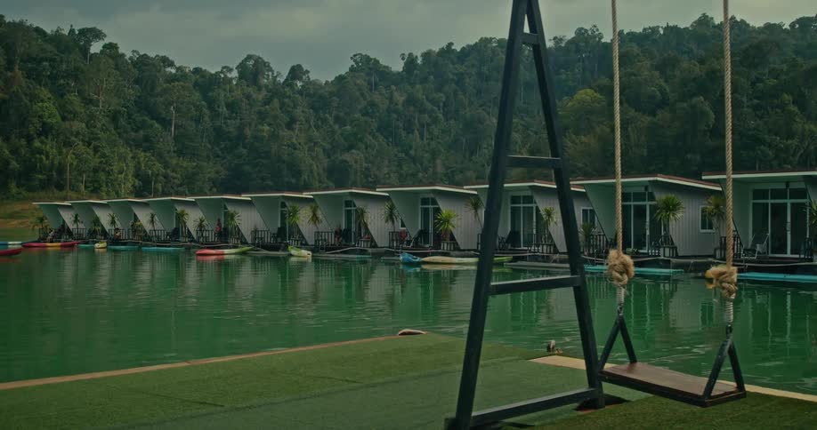 A tranquil view of a lakeside resort featuring a swing set and a row of floating cabins against a backdrop of forested hills