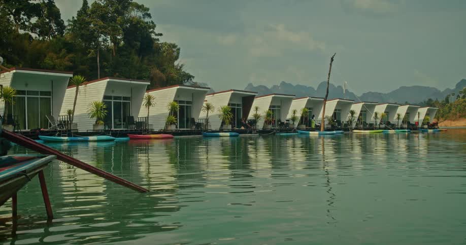 A traditional long-tail boat is moored near a row of modern floating bungalows at a serene lakeside resort nestled among mountains