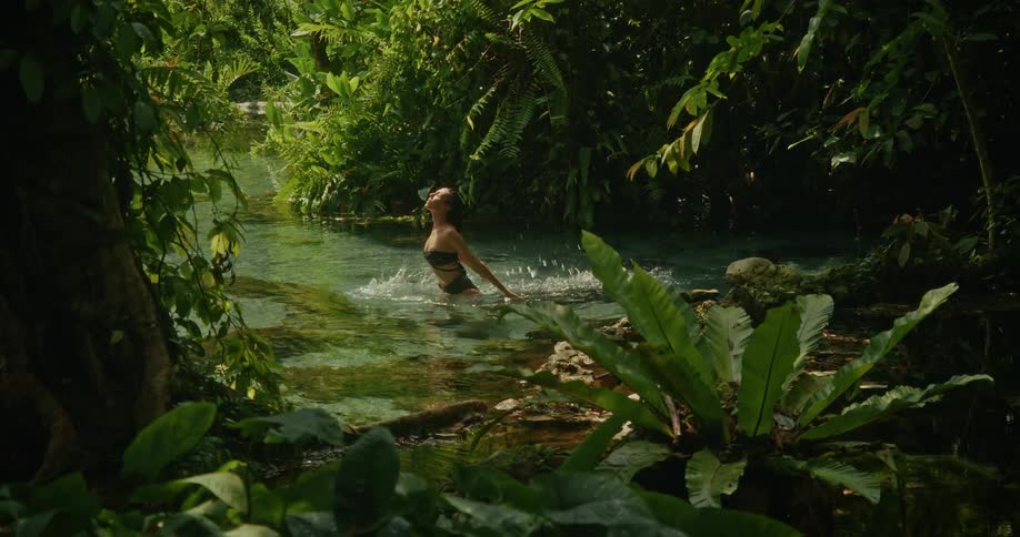 A woman in a black bikini flips her wet hair back in slow motion while bathing in a crystal clear river in a lush tropical jungle