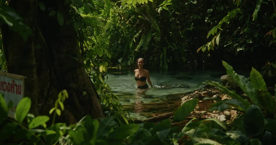 A serene woman in a black bikini wades through a clear jungle river surrounded by vibrant green tropical plants