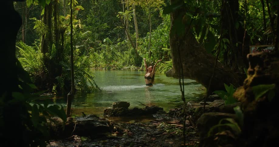 A slow-motion tracking shot reveals a happy woman in a bikini splashing in the clear water of a secluded jungle spring