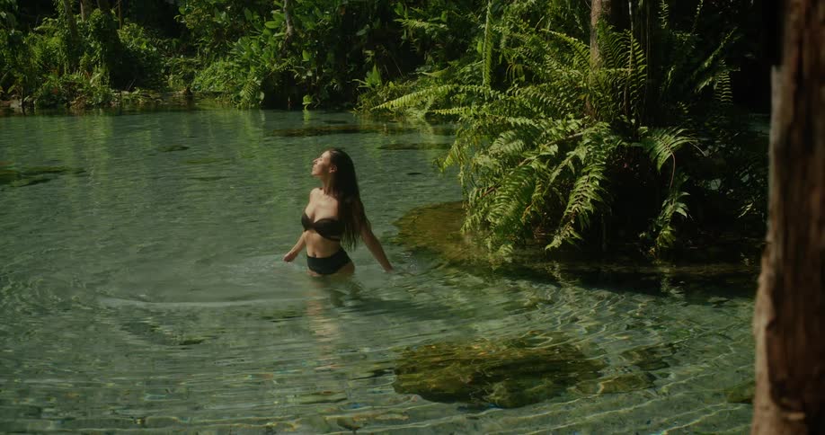 A beautiful woman in a black bikini enjoys the serene atmosphere while wading in a crystal-clear jungle spring