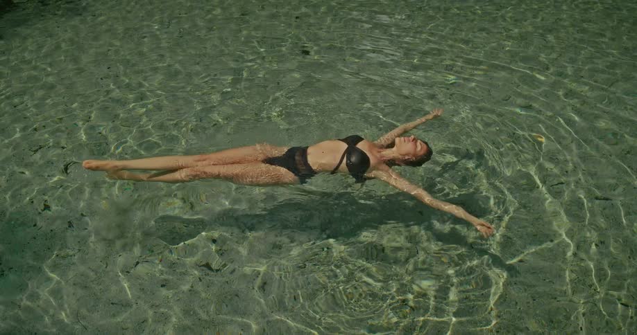 A serene woman in a black swimsuit gracefully floats on her back in the calm, crystal-clear water, basking in the sunlight