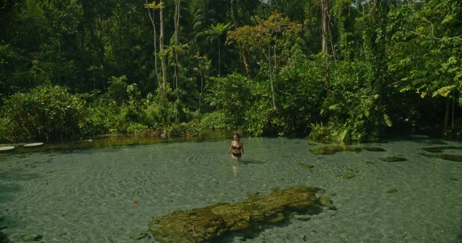 A serene woman in a bikini slowly walks through the crystal-clear water of a beautiful, secluded jungle spring