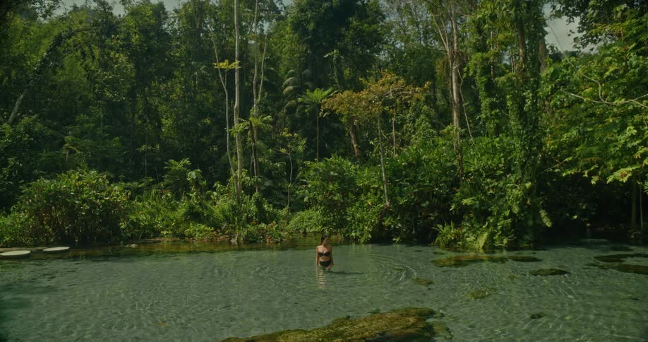 A woman in a bikini wades through a crystal-clear jungle spring, splashing playfully in the tranquil water surrounded by lush tropical foliage