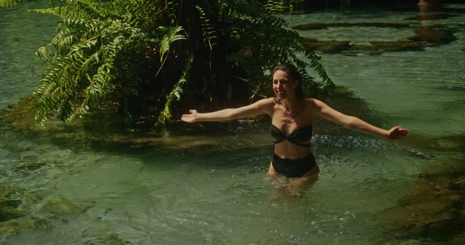 Slow motion shot of a joyful woman in a bikini splashing and swimming in a pristine, crystal-clear jungle river