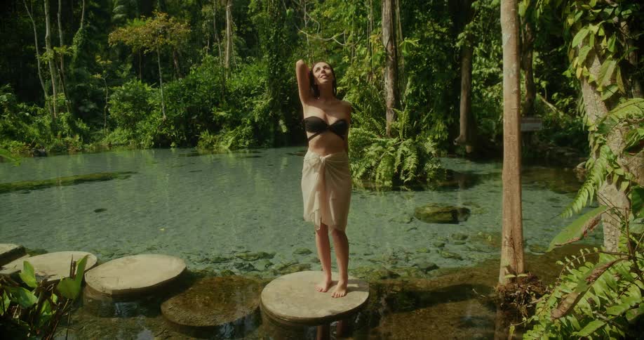 A serene woman walks across stepping stones in a crystal-clear jungle spring, stopping to feel the warmth of the sun in the tranquil rainforest