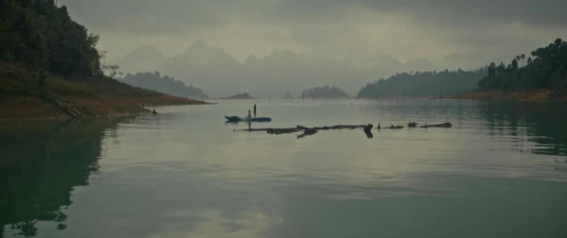 Camera tilts up from the water's surface to reveal a wide, serene view of a kayaker on a misty lake surrounded by mountains