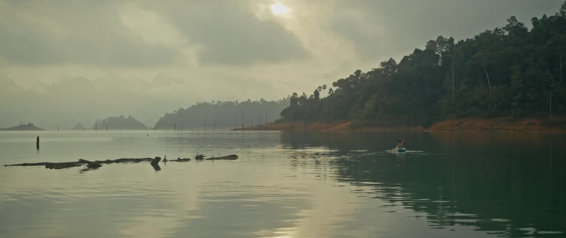 A tranquil wide shot of a kayak paddling across a vast, serene lake surrounded by a misty jungle at dawn