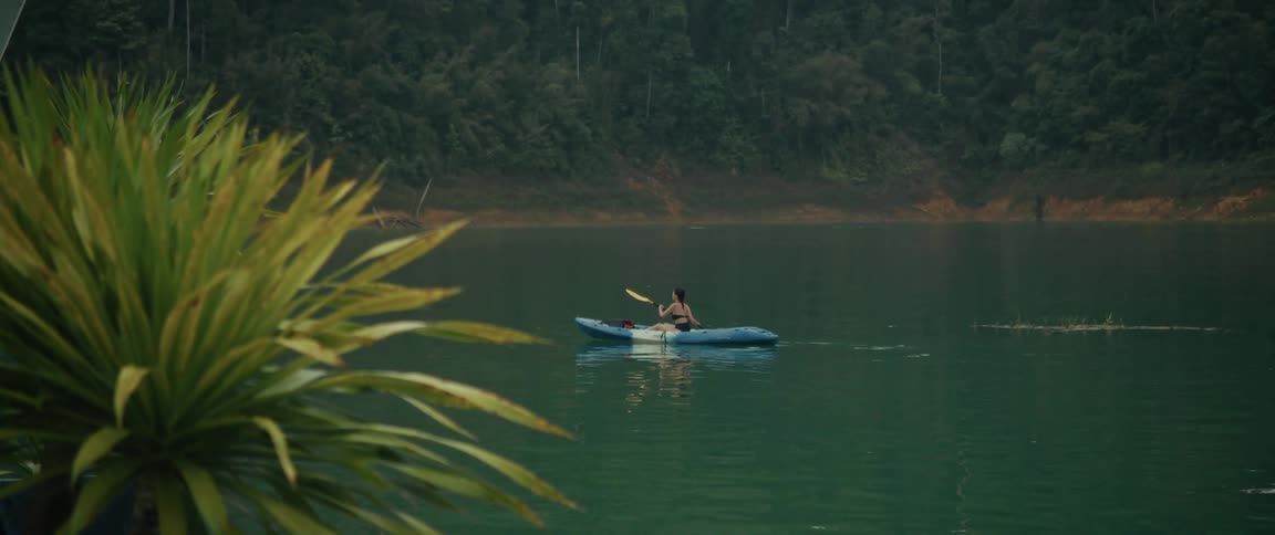 A woman in a kayak slowly paddles across a tranquil green lake, with a dense, misty forest along the shoreline