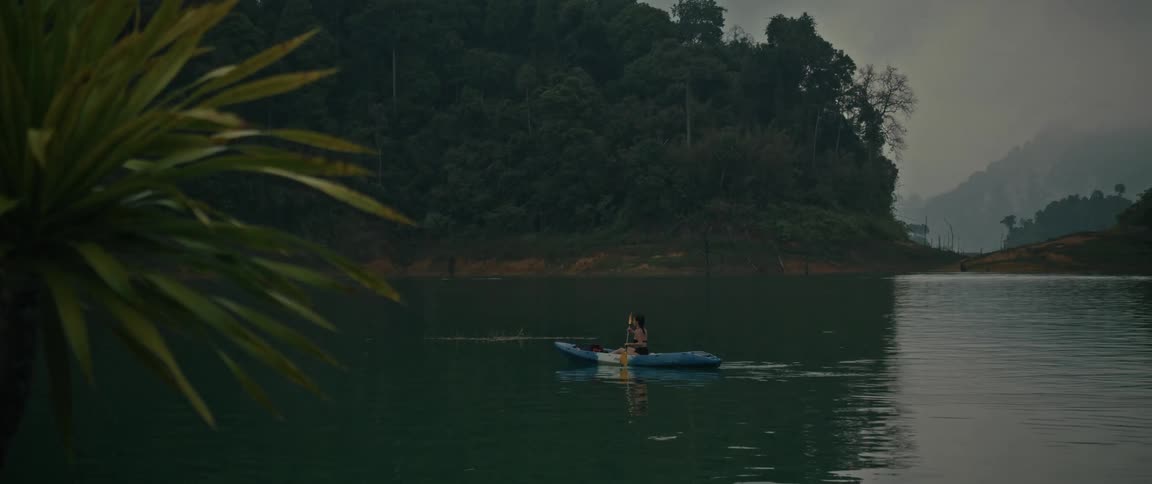 A lone kayaker peacefully paddles across a serene lake surrounded by a lush green forest on a misty morning