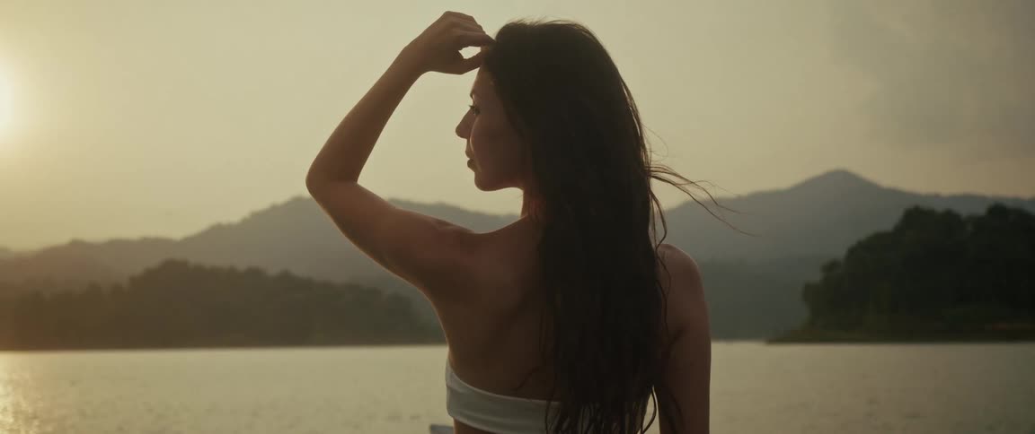 A woman with her back to the camera looks out over a tranquil mountain lake at sunset, her hair blowing in the wind