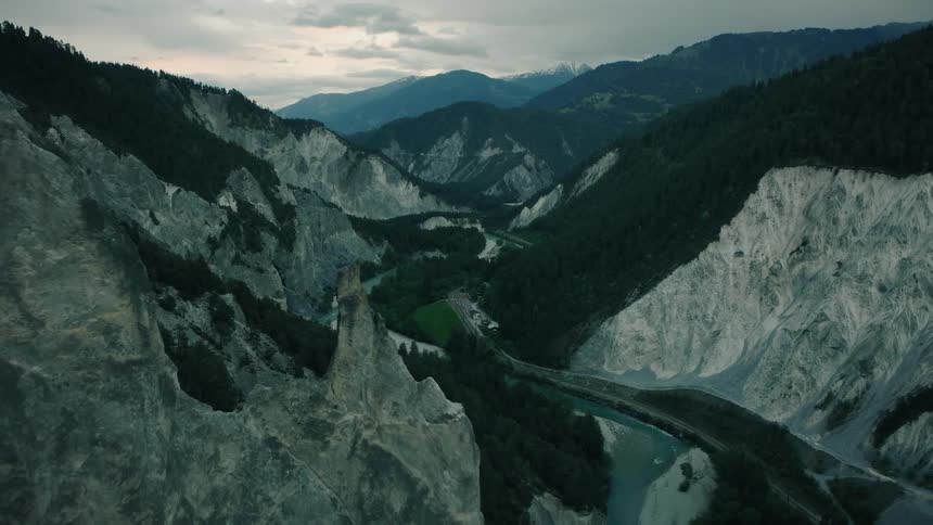 A dramatic aerial shot pulls back to reveal a jagged rock pinnacle overlooking a winding river in a vast mountain gorge