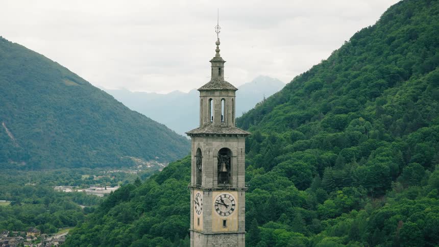 An old European clock tower is framed by lush green mountains under an overcast sky