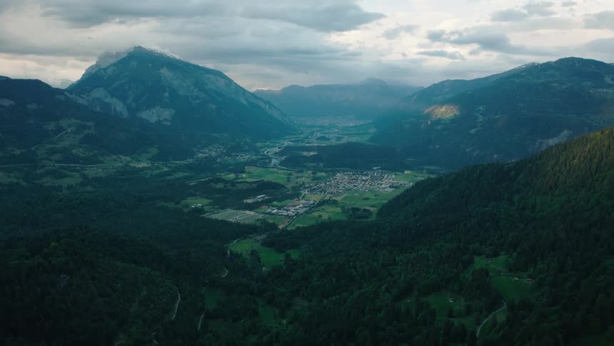 A majestic aerial view of a verdant valley with a small town nestled between towering mountains under a dramatic, cloudy sky