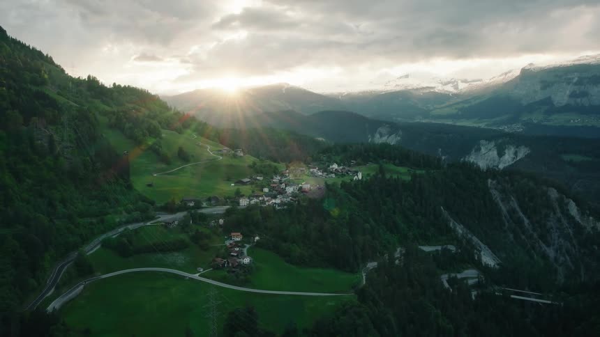 An epic aerial view of a small mountain village in a green valley as the sun sets behind snow-capped peaks