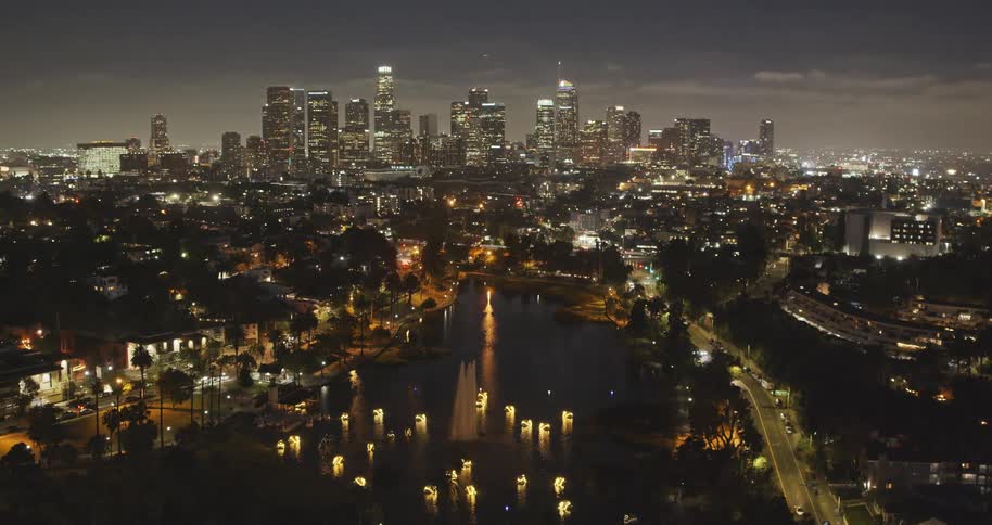 A stunning nighttime aerial view of the Downtown Los Angeles skyline, with the glowing fountains and swan boats of Echo Park Lake in the foreground