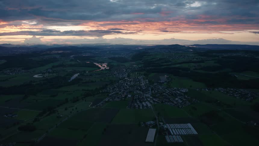 A dramatic aerial view shows a beautiful sunset casting a warm glow over a small town nestled in the rolling green hills of the countryside