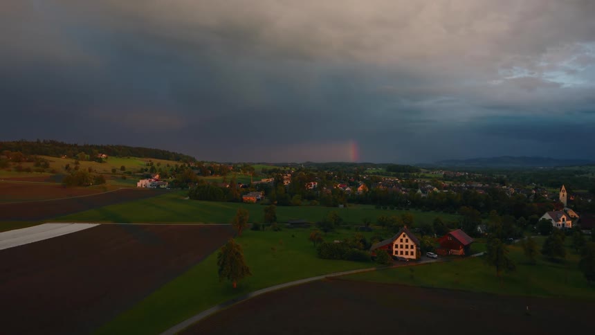 A dramatic aerial view shows a tranquil countryside village illuminated by sunset light under dark storm clouds with a faint rainbow