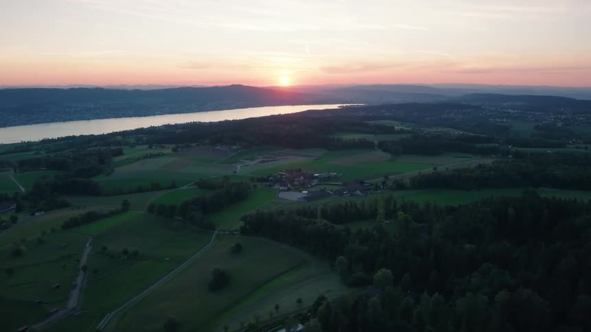 A serene, static aerial shot captures the beautiful sunset over a vast landscape of green fields, forests, and a large lake in the distance