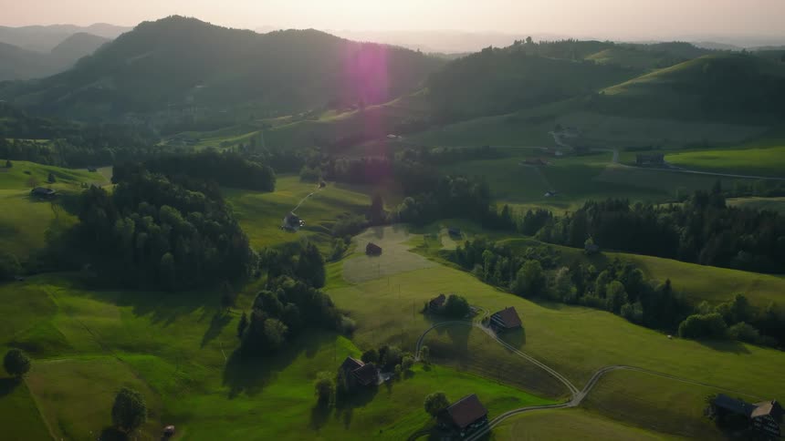 Stunning aerial view slowly pulling back over the idyllic rolling green hills and farms of the Swiss countryside at sunset