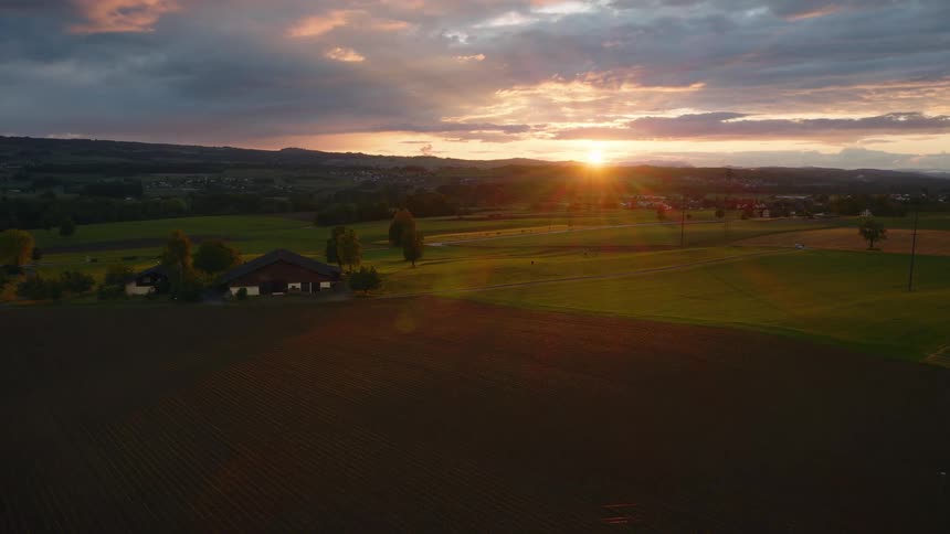 A static aerial shot captures a breathtaking sunset over a tranquil countryside landscape, with golden light bathing the fields and a distant farmhouse