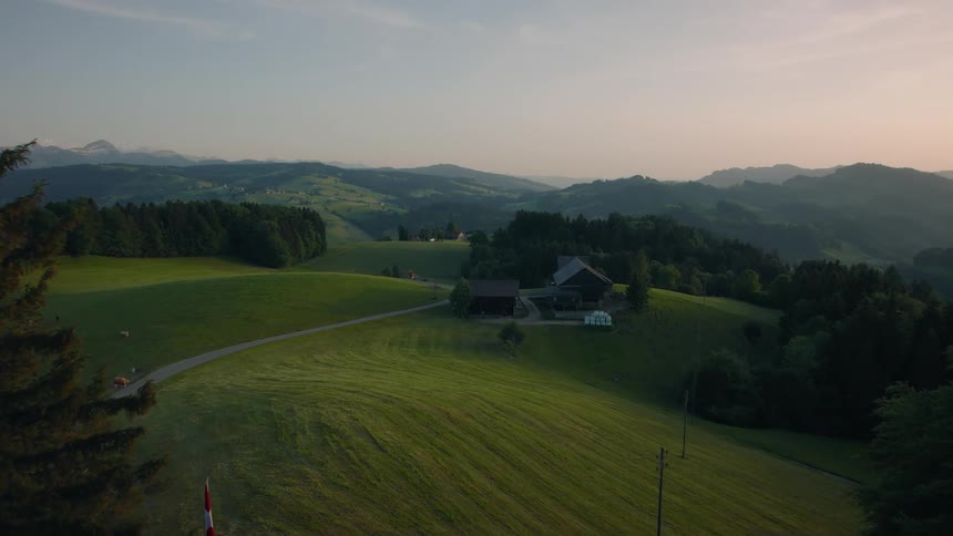 An aerial view reveals an idyllic Swiss farmhouse nestled in the verdant rolling hills of the countryside at sunrise
