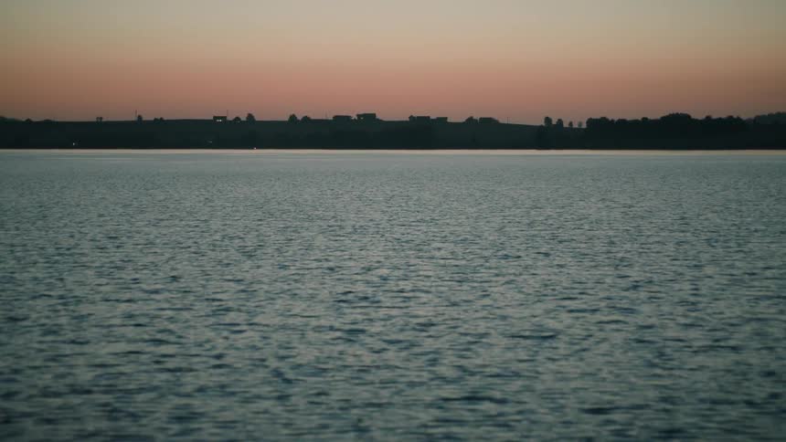 A serene pull-focus reveals the gentle ripples on a calm lake as the sun sets on the horizon