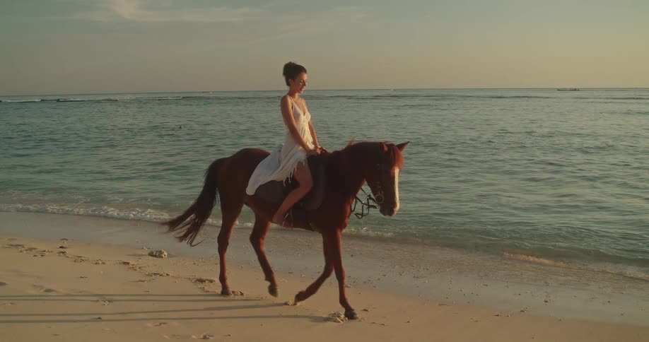 A beautiful woman in a white dress gracefully rides a horse along a sandy beach at sunset in slow motion