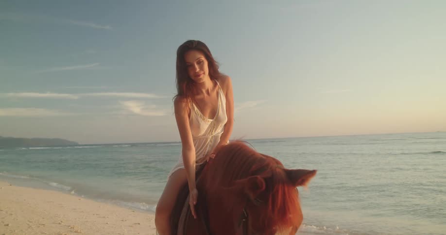A beautiful woman in a white dress sits atop a brown horse on a serene beach, smiling warmly as the golden sun sets over the ocean