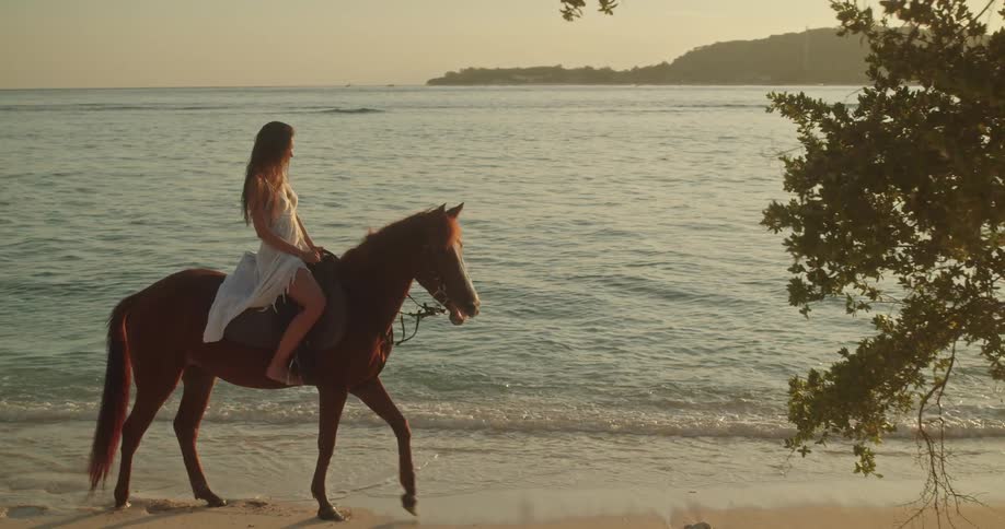A woman in a flowing white dress rides a horse along the ocean shore as the golden sun sets