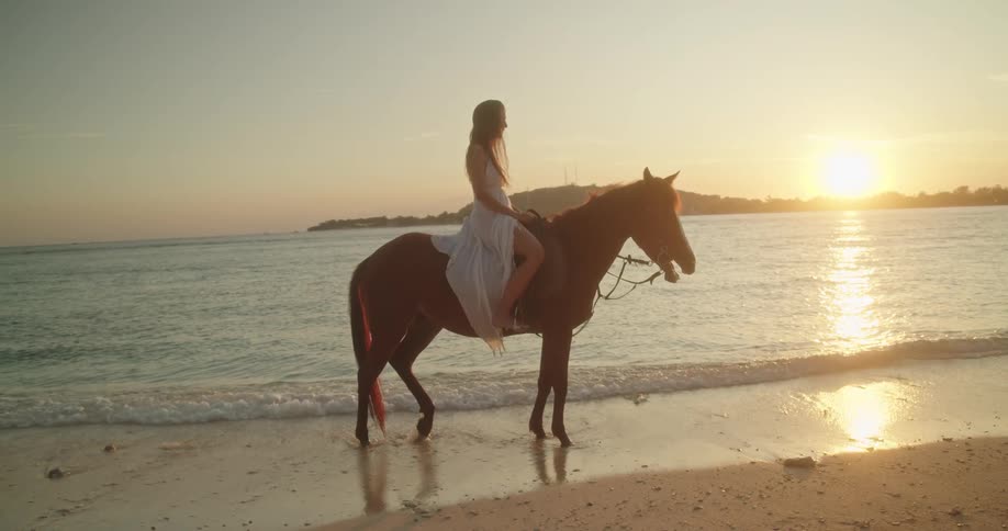 A woman in a beautiful white dress gracefully rides a horse on a sandy beach at sunset