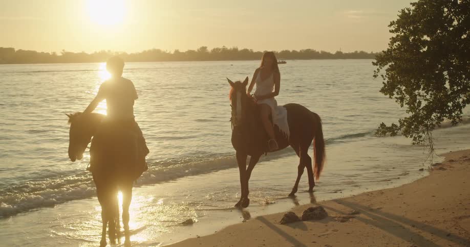 A woman in a white dress rides a horse along the ocean shore during a beautiful golden sunset