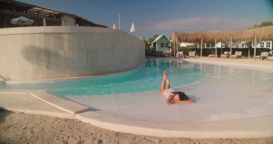 A woman in a white swimsuit relaxes in the shallow end of a unique, sand-edged swimming pool at a luxurious tropical resort