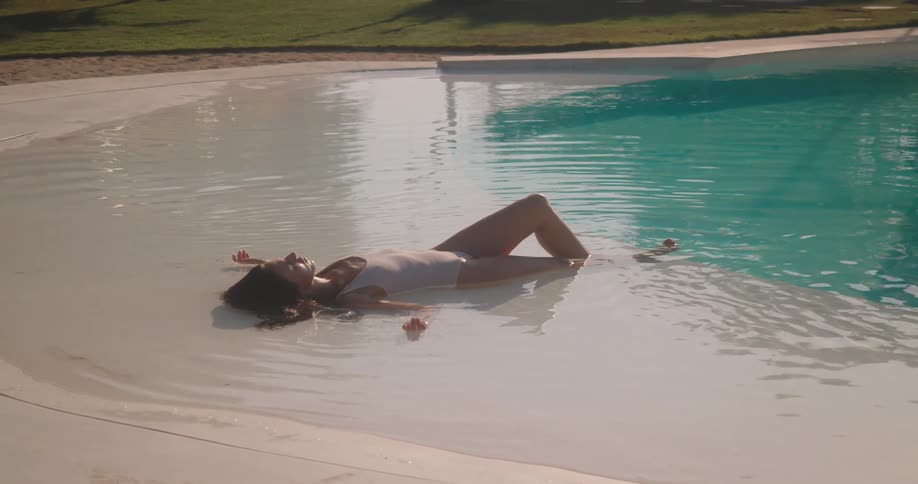 A woman in a white swimsuit peacefully relaxes in the shallow, sun-drenched waters of a luxury resort pool as the camera slowly zooms out