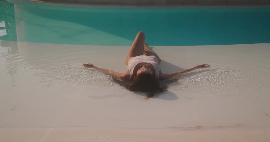 High angle shot of a woman in a white one-piece swimsuit relaxing in the shallow water of a luxurious, modern swimming pool