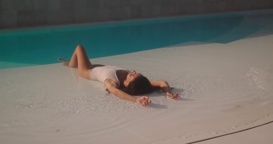 A slow-motion zoom-out shot reveals a beautiful woman in a white swimsuit relaxing in the shallow end of a modern swimming pool