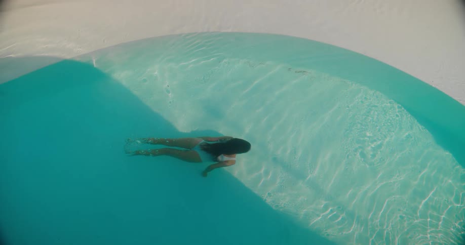A woman in a white swimsuit gracefully swims underwater and floats on her back in a minimalist turquoise pool, viewed from directly above in slow motion
