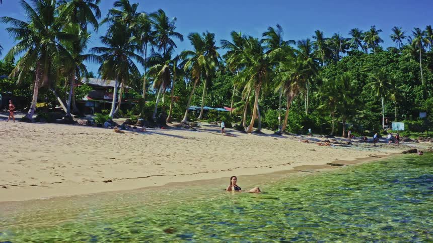 A woman relaxes in the crystal-clear shallow waters of a serene tropical beach lined with palm trees on a sunny day