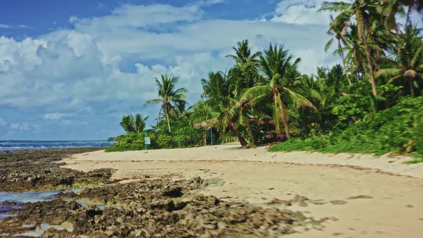 An aerial view flies slowly along a stunning tropical beach with white sand, palm trees, and a rocky shoreline under a beautiful cloudy sky