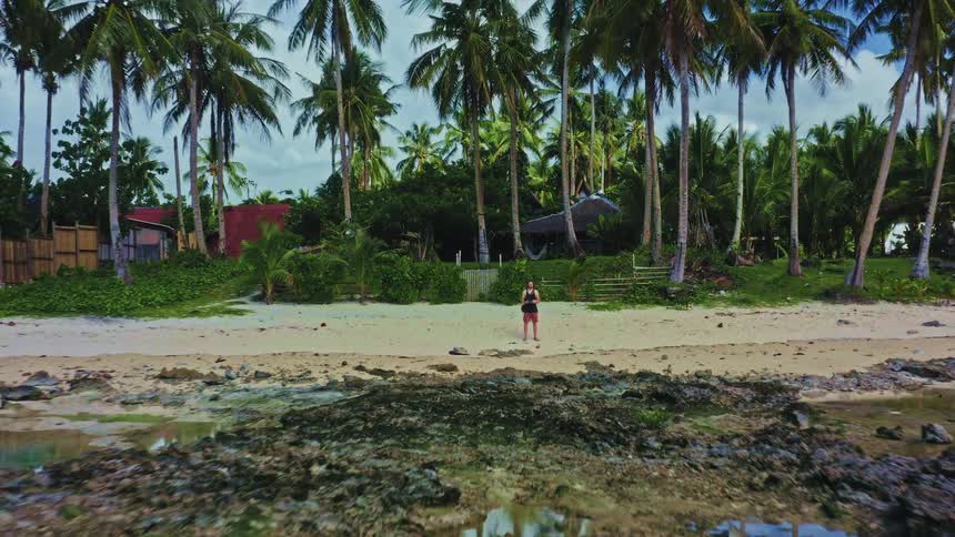 A male drone pilot waves at the camera as it pulls back to reveal a stunning tropical beach lined with palm trees