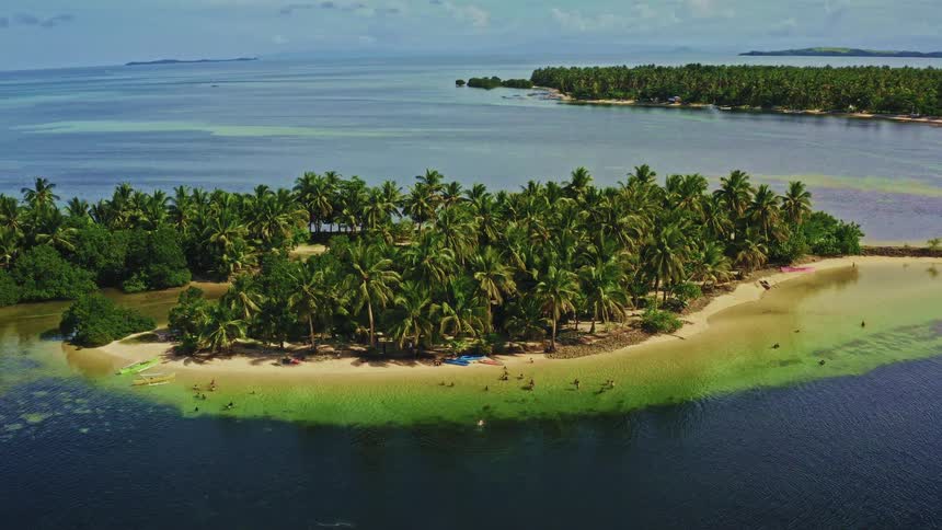 A stunning aerial view shows a small tropical island covered in palm trees where people are swimming in the shallow, clear water on a beautiful day