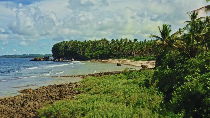 A slow aerial journey over a rugged, rocky shoreline towards a secluded tropical beach lined with lush palm trees