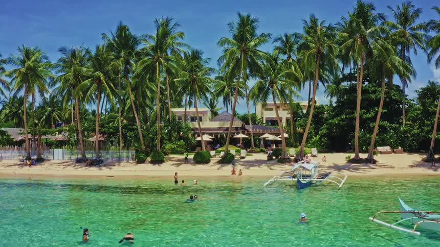 A slow aerial zoom-in captures the idyllic scene of a tropical beach resort, where people swim in turquoise waters and relax on the sand beneath swaying palm trees