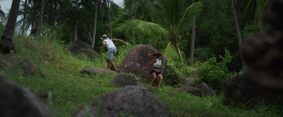 A man and a woman frantically escape through a rocky jungle landscape in slow motion
