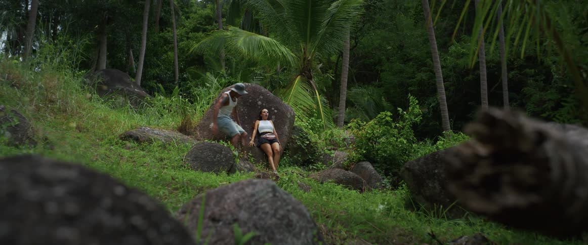 A playful couple runs and jumps through a lush tropical jungle in slow motion before stopping to rest on a boulder