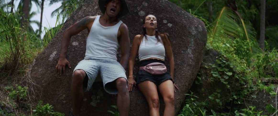 A terrified man and woman run for their lives through a dense jungle, finally collapsing in exhaustion and fear against a large boulder