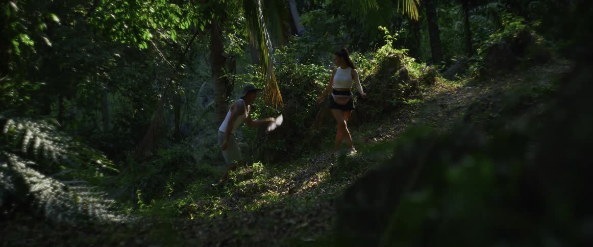 A man fans his partner with a hand fan to cool her down as they hike together through a lush, tropical jungle