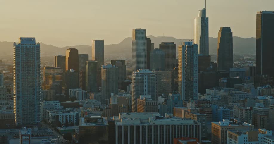 An aerial view of the Downtown Los Angeles skyline with its iconic skyscrapers bathed in the warm, hazy light of a golden hour sunset